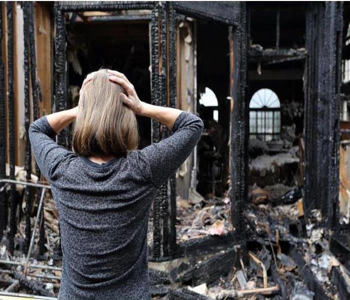 woman standing in front of a home after a fire has caused damage with both hands up holding her head.
